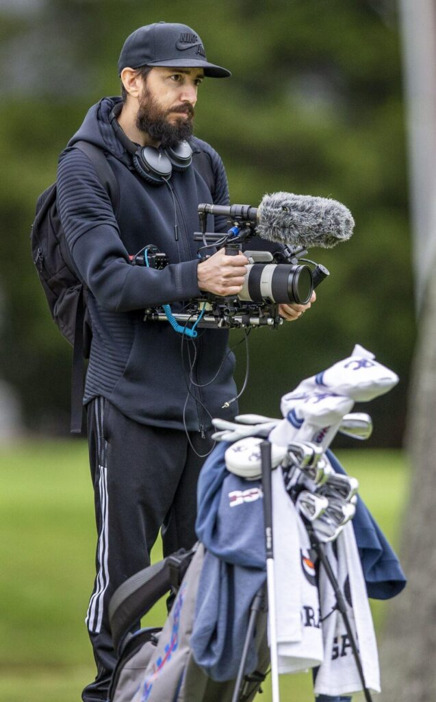 with a bag of golf clubs in the foreground, a man stands holding a high tech camera