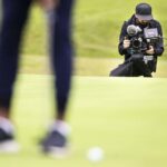 on a golf green, a man kneels with a camera, filming some one playing golf