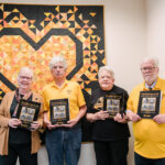 Four alumni hold books in front of a large quilt.
