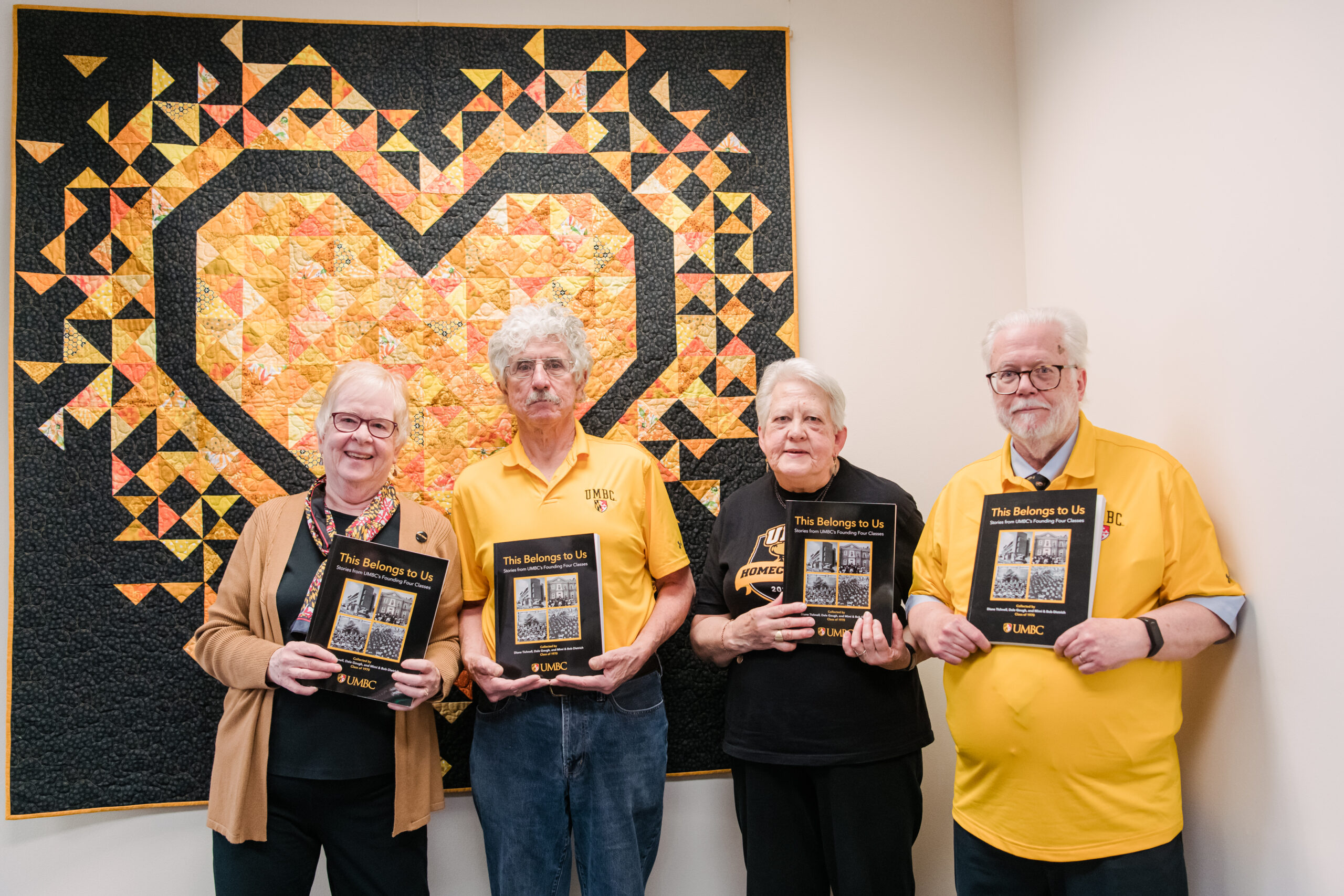 Four alumni hold books in front of a large quilt.