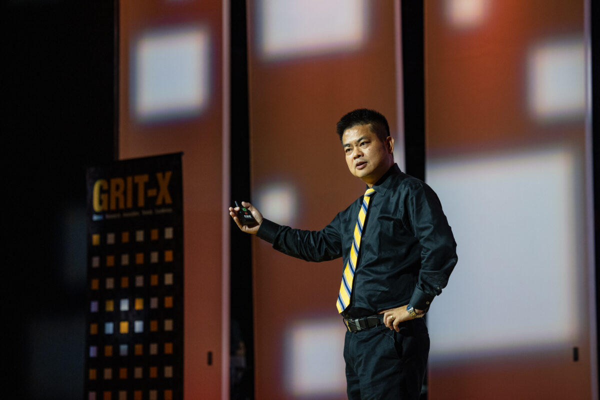 Zhensen Huang on stage delivering a talk. He is holding out his left arm and in his left hand is a remote. 