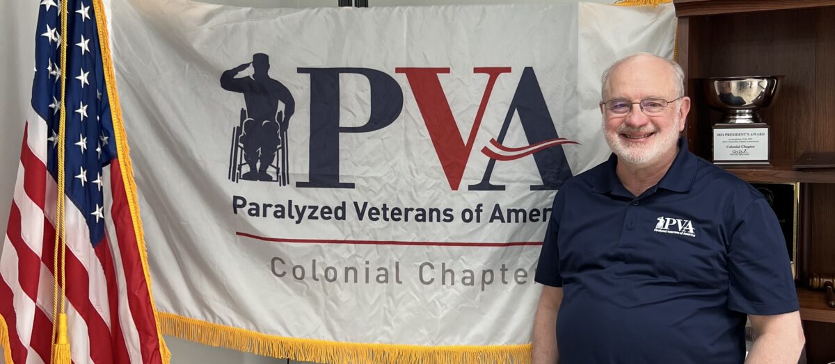 an older man stands in front of a flag that says Paralyzed Veterans of America