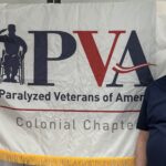 an older man stands in front of a flag that says Paralyzed Veterans of America