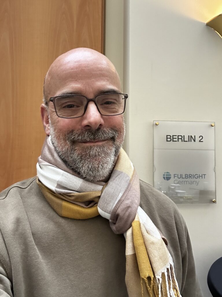 a man in a yellow and grey scarf smiles in front of a sign that says Fulbright, a study abroad exchange program