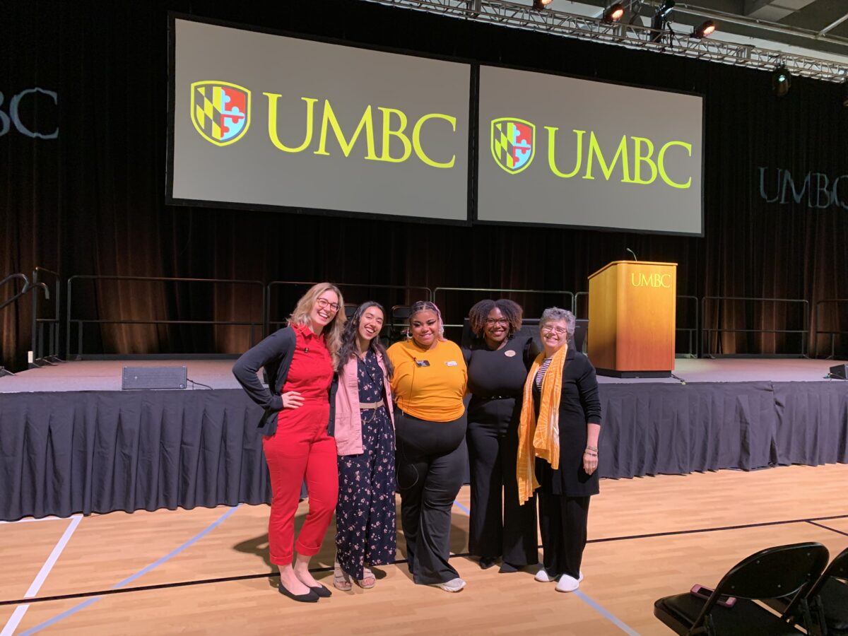 A group of women stand in front a stage and screens that say UMBC. They are members of the group Your Story Belongs Here. 