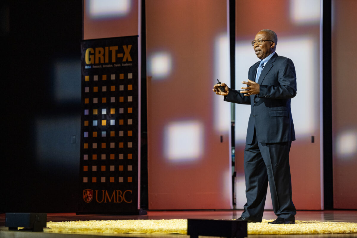 Charles Ichoku on stage delivering his GRIT-X presentation. He is gesturing with his hands. 