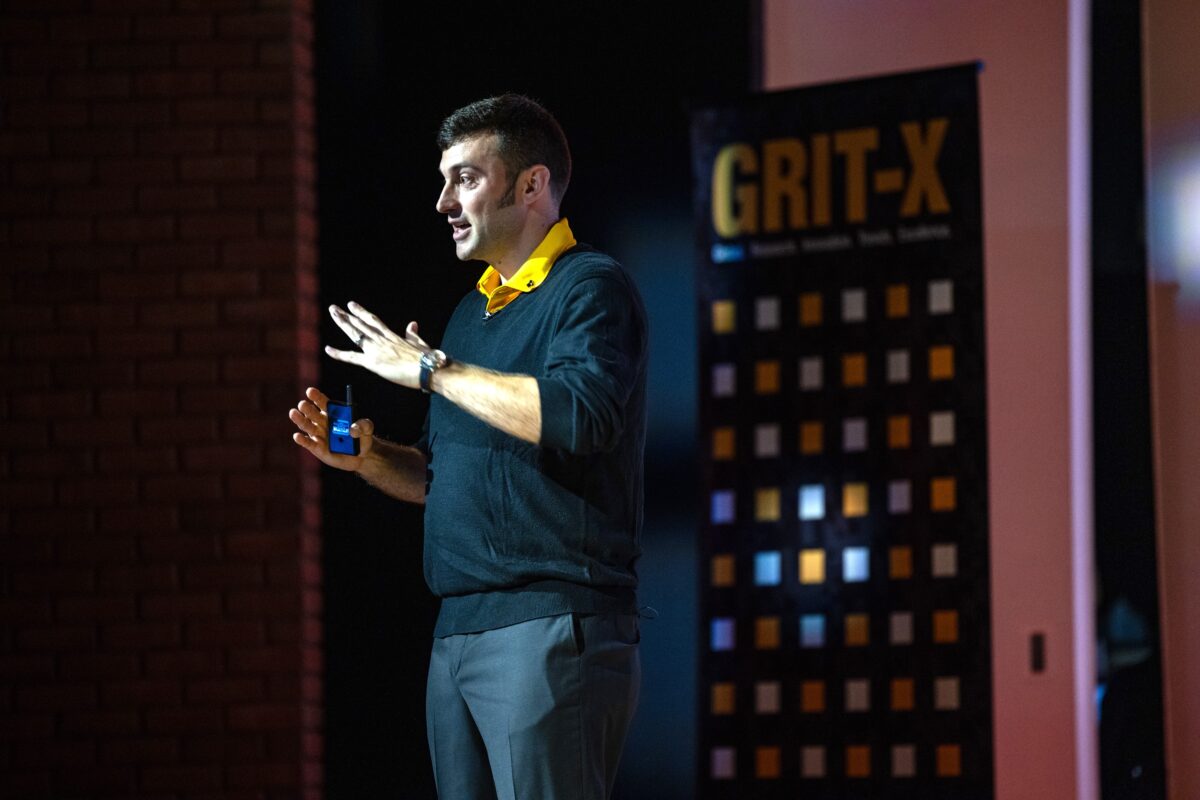 Justin Webster delivering his GRIT-X 2023 talk on stage at UMBC. He is gesturing with his hands and has a remote in his left hand.