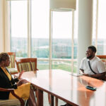 A woman and a college student sit talking to each other across a conference table with lots of windows behind them during office hours