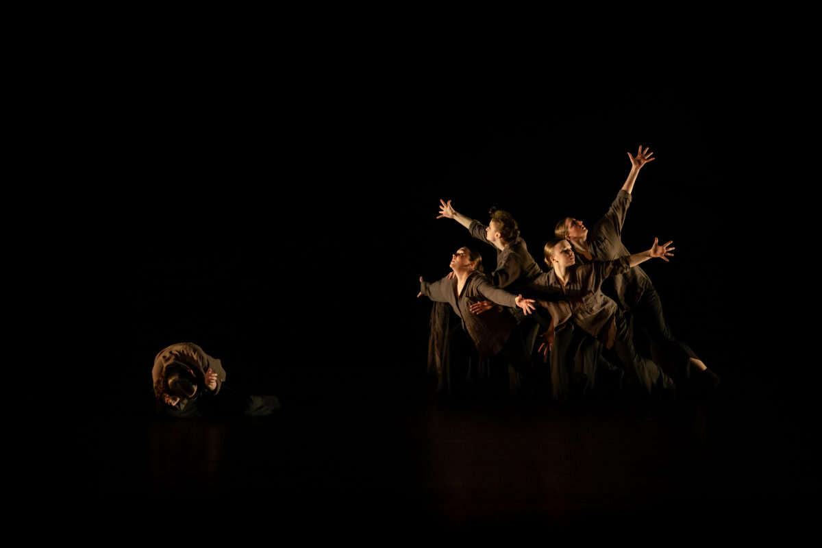 A group of dancers in dark tunics thrust their arms in the air on a stage
