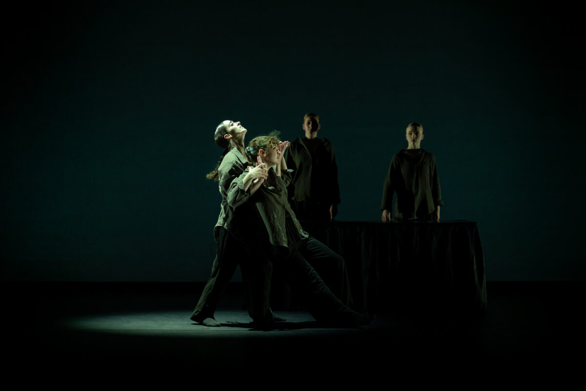 Dancers on a darkly-lit stage hold another dancer up