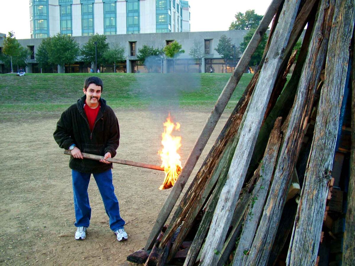 Thomas Locastro holds a flaming stick to an unlit bonfire in the foreground and smiles.