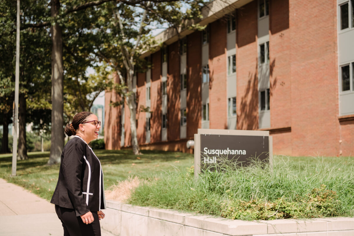 A woman in a black business suit stands next to a university dorm