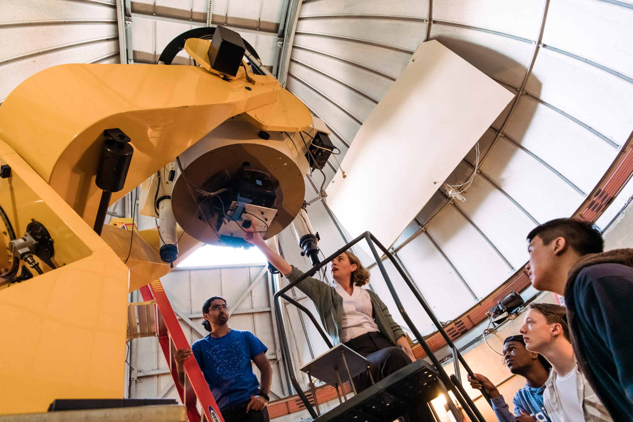 woman in center standing on stepladder, reaching upward toward a large lens held by a huge yellow frame. Four students around her look up at what she's doing; one is standing on a ladder.