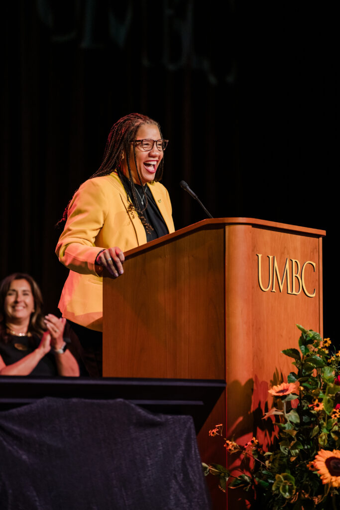 a woman in a gold blazer speaks at a lectern that says UMBC