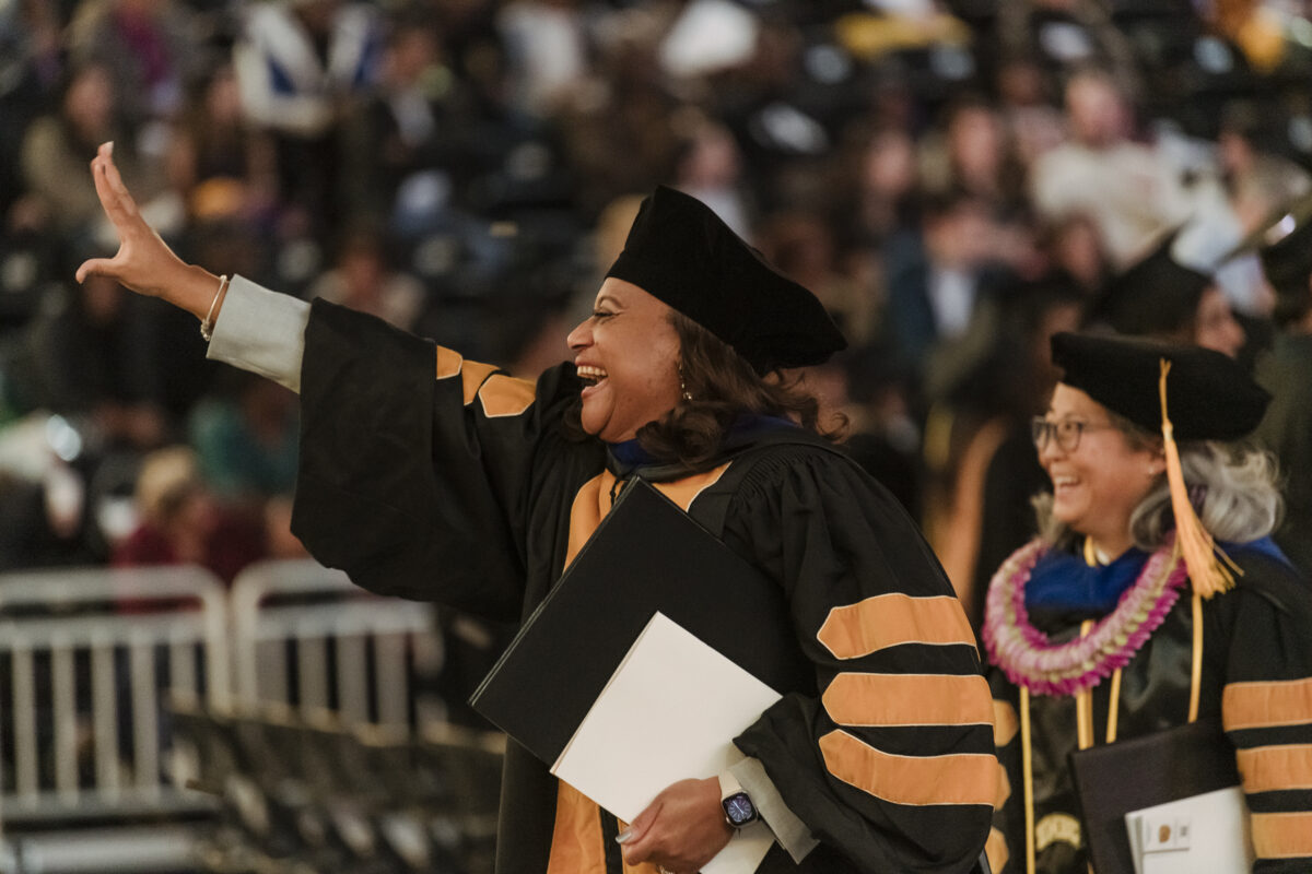 A Ph.D. graduate smiles with hand outstretched in a wave