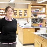 portrait of woman wearing a black sweater in a laboratory, backed by lab counters and shelving with chemicals and flasks
