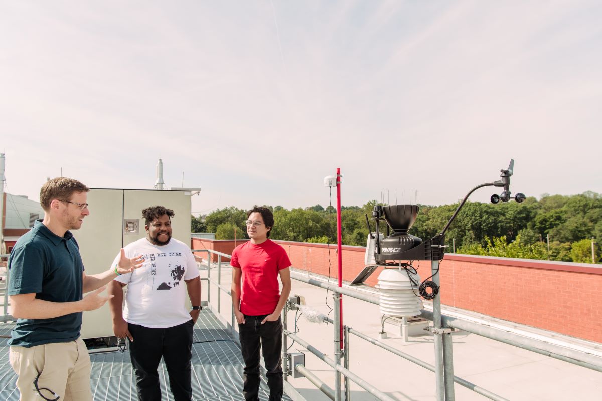Three people stand on a roof next to equipment. Trees in distance.