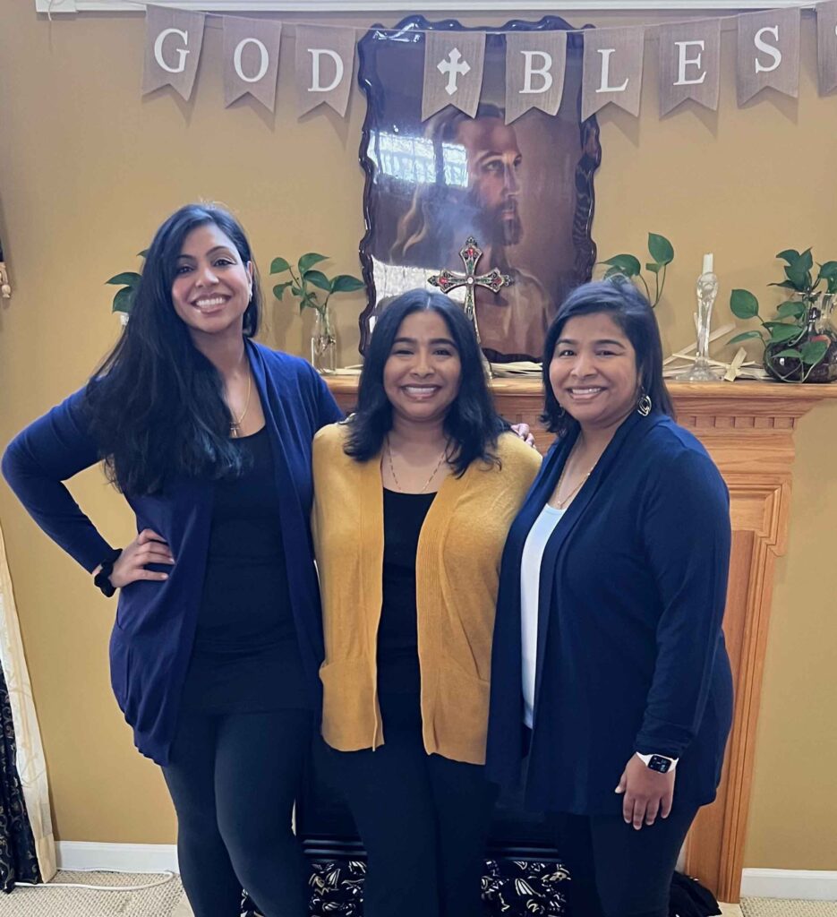 three adult sisters pose in front of a sign that says God Bless