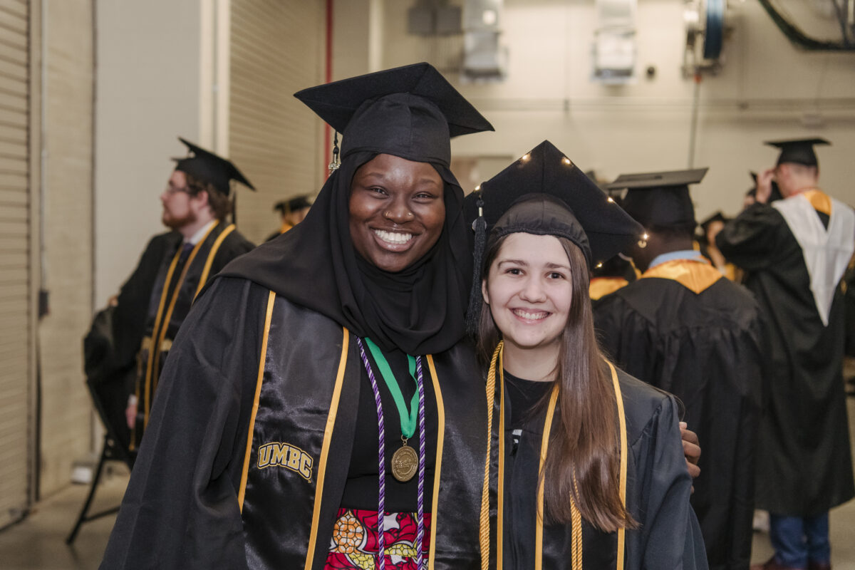 Two students in commencement regalia and robes stand smiling next to each other