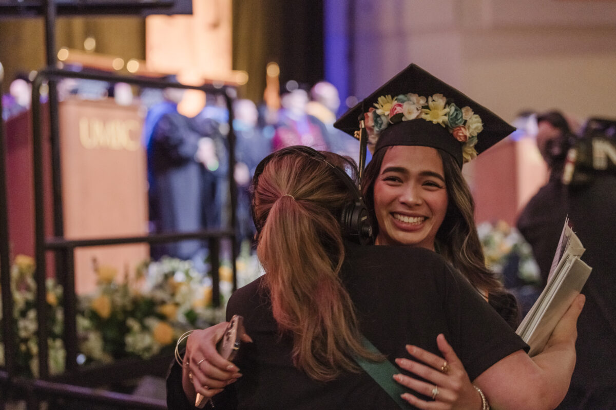 A student wearing a mortarboard with flowers hugs a staff member