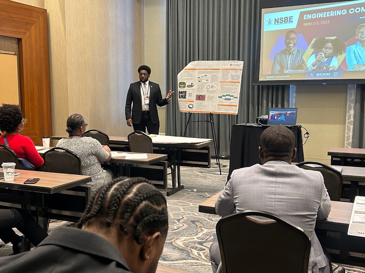 man speaking standing next to a research poster with a screen behind him that reads "NSBE Engineering Conference, Nov 3- 5, 2023"; seated audience members listen