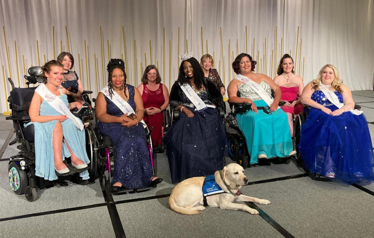 five women in ballgowns pose in wheelchairs wearing sashes. a guide dog lays down in the front.