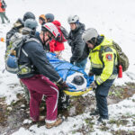 A group of people wearing winter gear and helmets carry a stretcher with a fake body. disaster health systems emergency