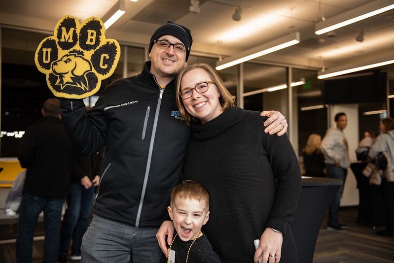 A family embraces and smiles at the camera. The father has a foam UMBC paw.