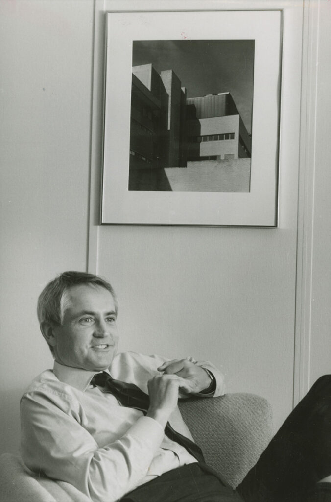 black and white portrait of Michael Hooker sitting in a chair below a framed photograph