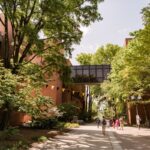 Students walk between brick academic buildings. Trees with leaves line walkway.