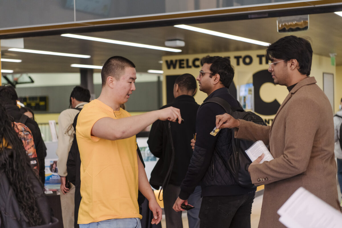 A person wearing a gold colored t-shirt holds a digital scanner for students to scan their plastic IDs 