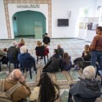 A group of people inside a mosque sit on a carpeted floor listening to The imam of the Khadija Mosque