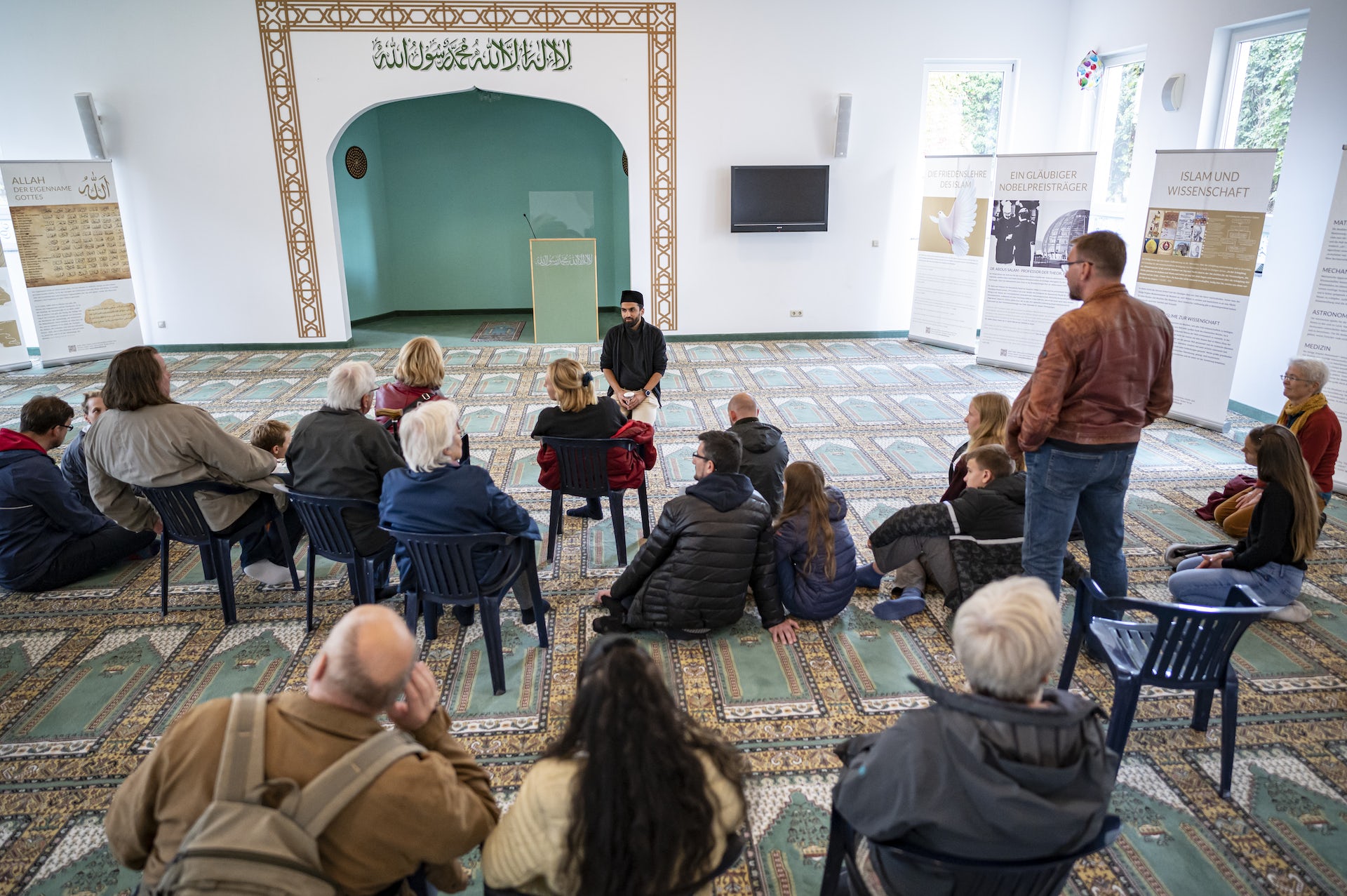 A group of people inside a mosque sit on a carpeted floor listening to The imam of the Khadija Mosque