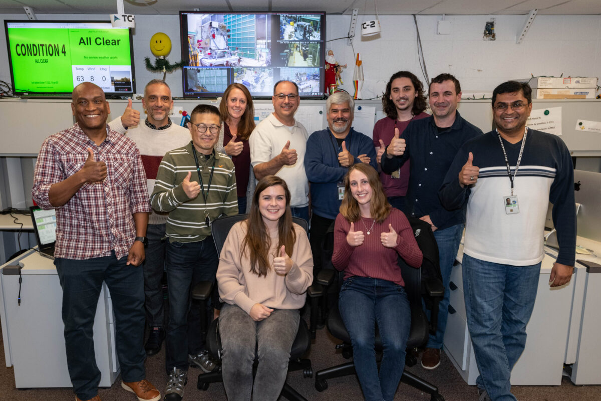 group photo, everyone giving a thumbs up. Several webcam scenes of the spacecraft on a screen behind the group; also a bright green screen that reads "All Clear"