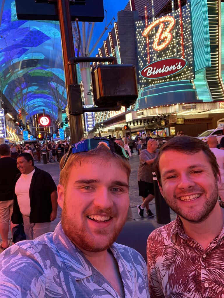 Two men (one is a Russian speaker) take a smiling selfie in front of a lot of lit up billboards in Las Vegas at a cybersecurity conference