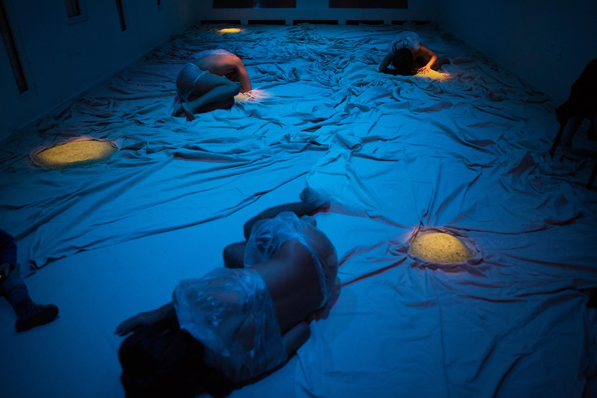 In a dimly lit scene bathed in blue, three dancers lie on a rumpled cloth floor