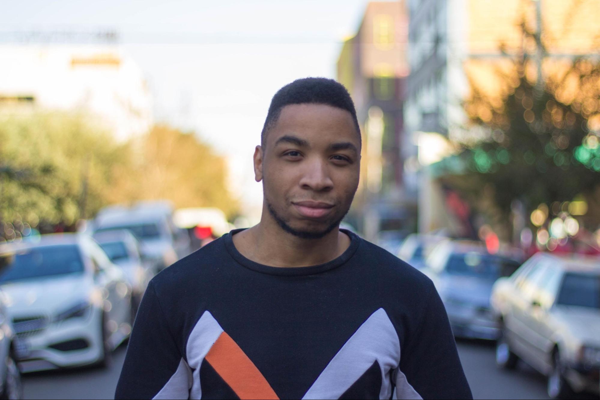 Against a blurry outdoor street scene, a Black man in a dark top smiles at the camera