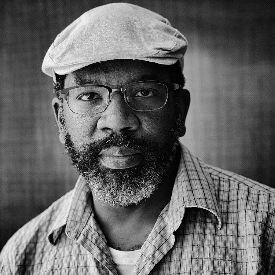 In a black and white image, a Black man with glasses, beard and a cap looks at the camera