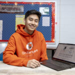 student in bright orange hoodie sits at desk in classroom next to laptop with research slides on screen