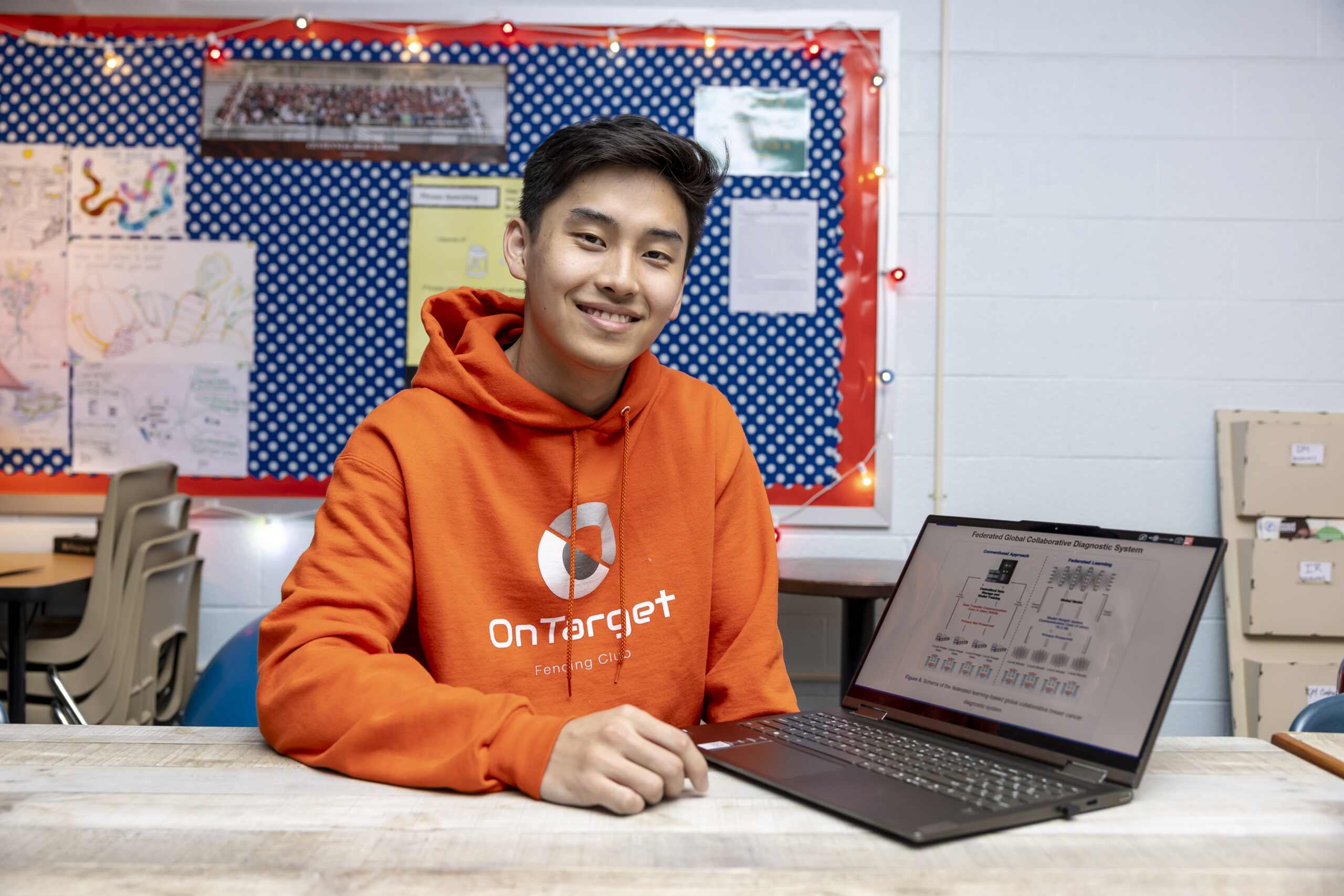 student in bright orange hoodie sits at desk in classroom next to laptop with research slides on screen