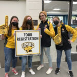 four students in UMBC black and gold attire stand behind a poster that reads "Tour Registration". Two hold yellow foam thumbs-ups.