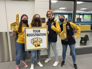 four students in UMBC black and gold attire stand behind a poster that reads "Tour Registration". Two hold yellow foam thumbs-ups.