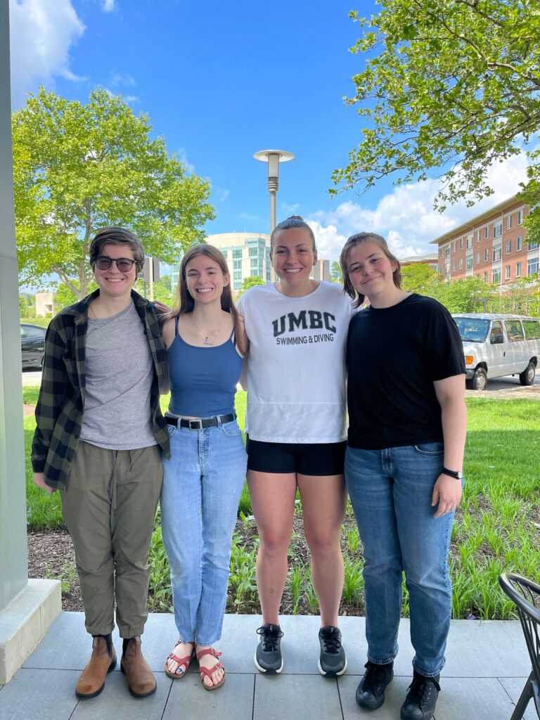 group photo of four students on the UMBC campus, spring greenery in the background