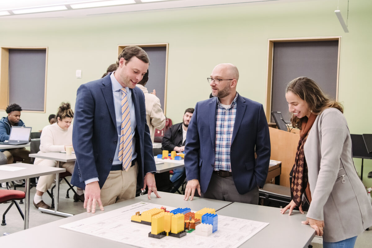 Three people dressed in business clothing stand around a table analyzing a LEGO-built urban development neighborhood.