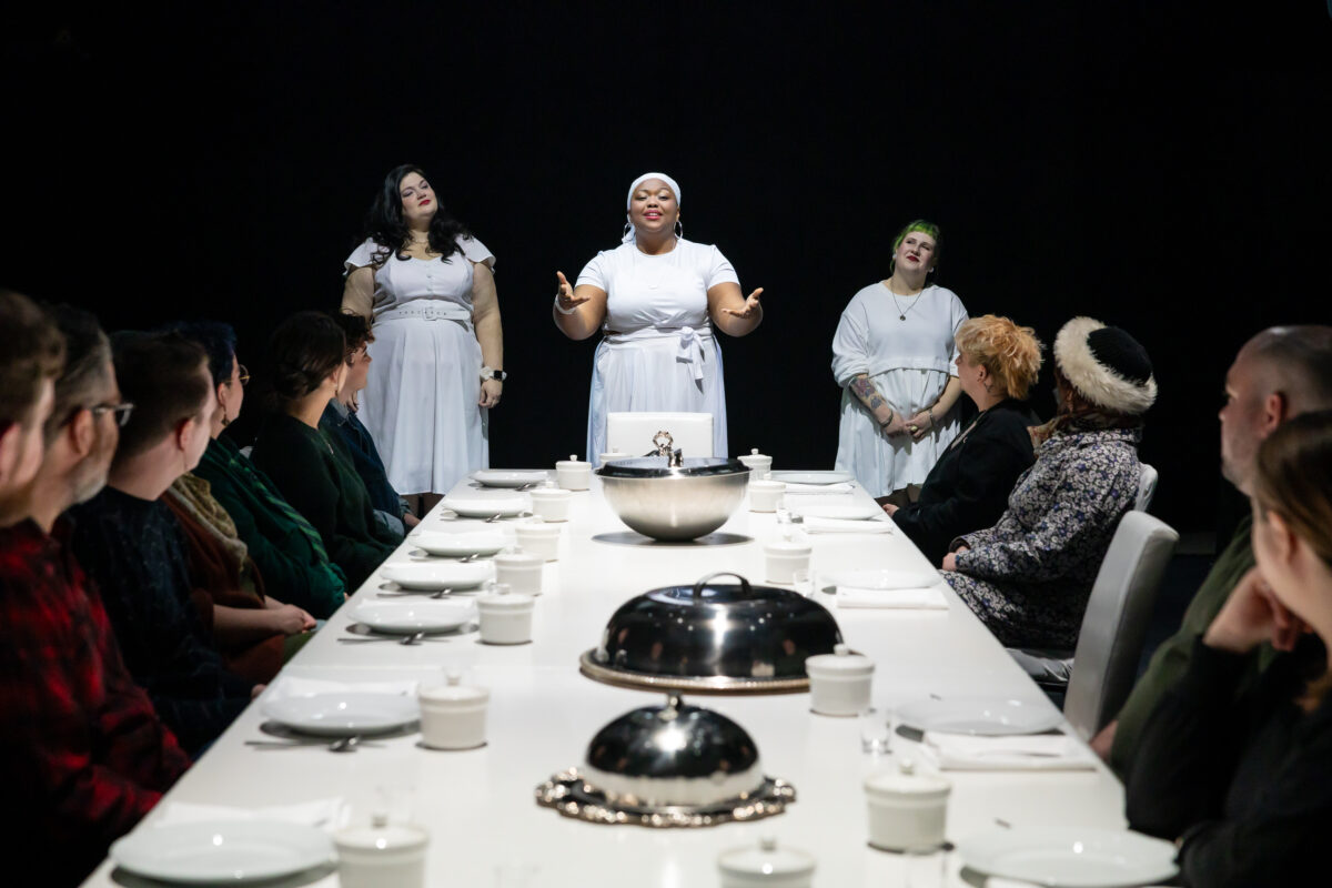 In a theatrical setting, we see a long dinner table covered with a white tablecloth. Three women stand at the head of the table.