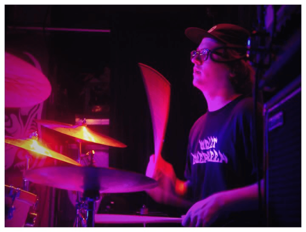Man sitting behind a drum kit playing under purple lighting.