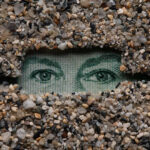 In a work of art, two eyes gaze out from a rectangular enclosure surrounded by small stones.