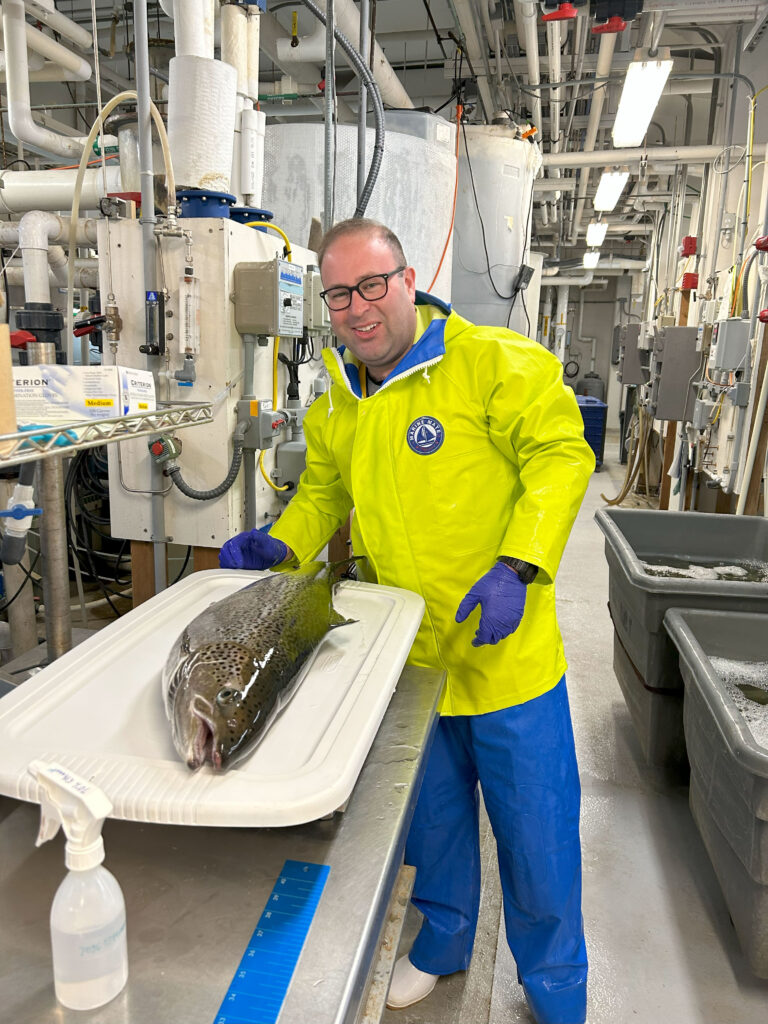 Jonas Miller stands wearing a safety yellow jacket, blue waterproof pants, and gloves next to a tray scale that holds a large fish. He is in a large research area with numerous tanks, bins, and pipes at IMET