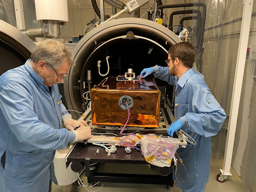 Two reseachers working on the LEMS lunar instrument. There are many wires and parts connected to a suitcase-sized instrument. 