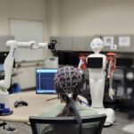 Woman wearing EEG cap sits with back toward camera. A computer screen, robotic arm and humanoid robot sit in front of her.